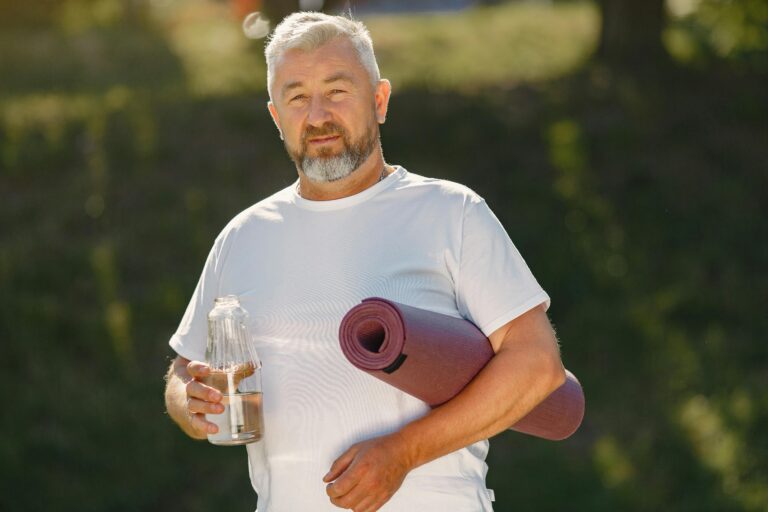 Senior man holding a yoga mat and water bottle outdoors, ready for exercise.